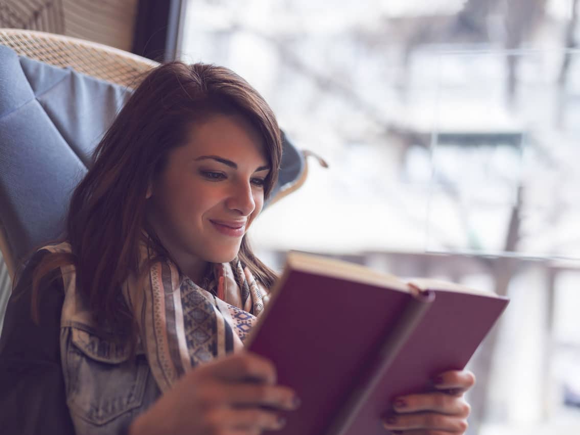 adobestock_168190634 Dieses Foto zeigt eine junge Frau, wie sie lächelnd und gedankenversunken in einem Buch liest. Sie hat dunkle lange Haare, trägt eine Jeansweste und ein blau-weiß gemustertes Halstuch. Sie sitzt am Fenster in einem Sessel. Das Bild soll zeigen, dass jeder von uns Trance kennt, wie beim Lesen eines Buches. Wie mit dem Flow in dem man sich beim Lesen befinden kann, ist es auch möglich mit Hypnose, Coaching Online und Psychotherapie Entspannung zu erfahren.